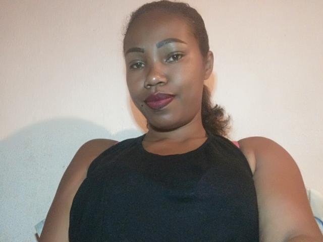 Tiane, com pele escura, olhos castanhos médios e cabelo preto em um rabo de cavalo, vestindo uma blusa preta sem mangas, mostrando seus seios e usando um piercing no nariz, com batom roxo..
