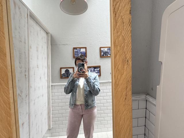 JaneRoa with a camera in a bathroom mirror, wearing a denim jacket, white shirt, and pink pants, standing in front of marble and white-tiled walls.