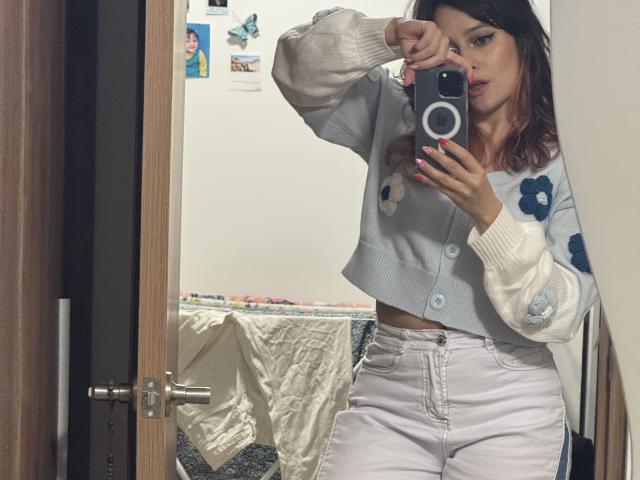JaneRoa with medium tits, wearing a white cardigan with blue flowers, white high-waisted pants, taking a mirror selfie, holding a camera, in a bedroom with a wooden door.