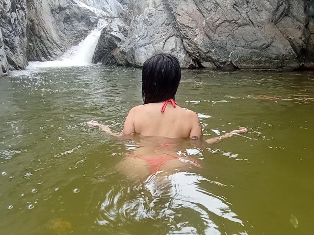 AlisonKweel, bare back, red bikini bottom, wet black hair, arms spread, in green water, facing waterfall, rocky walls. Her ass is visible, water splashes around.