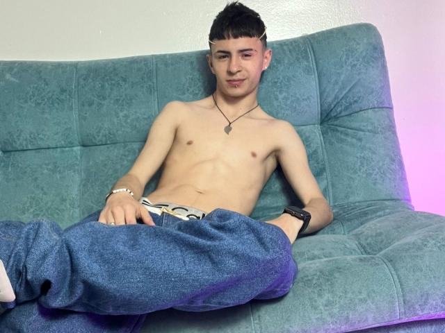 Shirtless StivarForte with a slim build, dark short hair, and a slight smirk, wearing blue pants and a necklace, sitting on a green couch, wristwatch on.