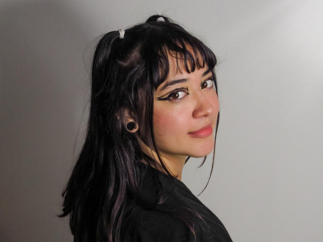 AzumiFujii with black hair, white hair ties, dark eyeliner, black ear plugs, and a subtle smile, wearing a black shirt, looking over her shoulder.