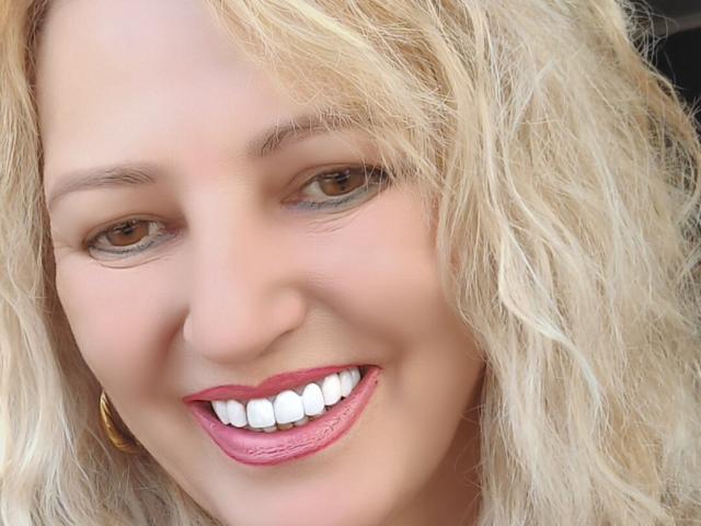Le visage de SexyCoco, cheveux blonds ondulés, yeux marrons, rouge à lèvres rose, dents blanches, sourire, boucle d'oreille dorée, peau claire, maquillage léger, aspect légèrement négligé, gros plan, attrayante..