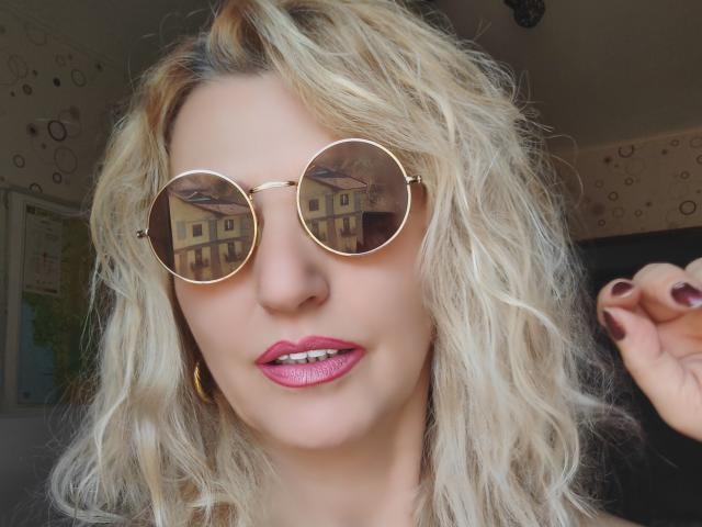 SexyCoco com cabelos loiros ondulados, usando óculos de sol redondos dourados, batom rosa brilhante e brincos de argola dourados, mostrando esmalte de unha marrom, com uma aparência fofa e elegante..