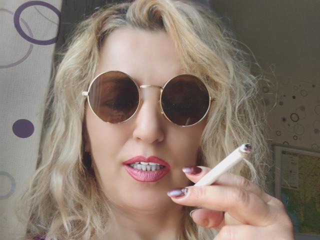 SexyCoco with blonde wavy hair, pink lipstick, round aviator sunglasses, holding a cigarette, pink manicured nails, and a slightly open mouth showing teeth.