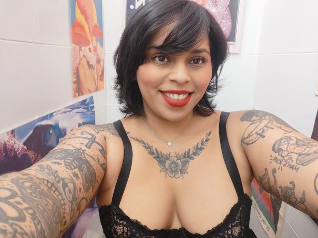 Chili, a tattooed woman with black hair and red lipstick, takes a selfie while smiling, wearing a black lace bra.