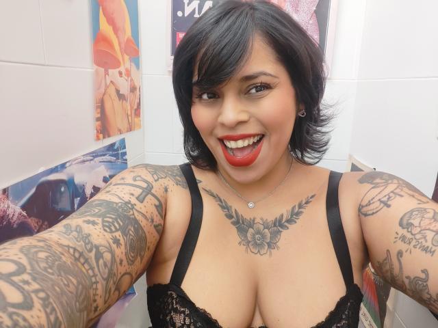 Chili with black hair and red lipstick is smiling, showing off her large breasts and detailed tattooed arms in black lace lingerie.