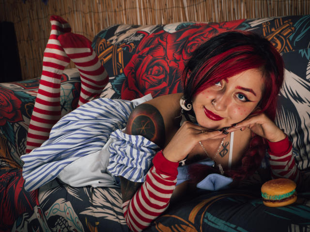 MeiJaisen with red and black hair, striped socks, and tattoos, lying on a rose-patterned couch, wearing a blue-striped dress, red arm sleeves, and a ring, with a donut.