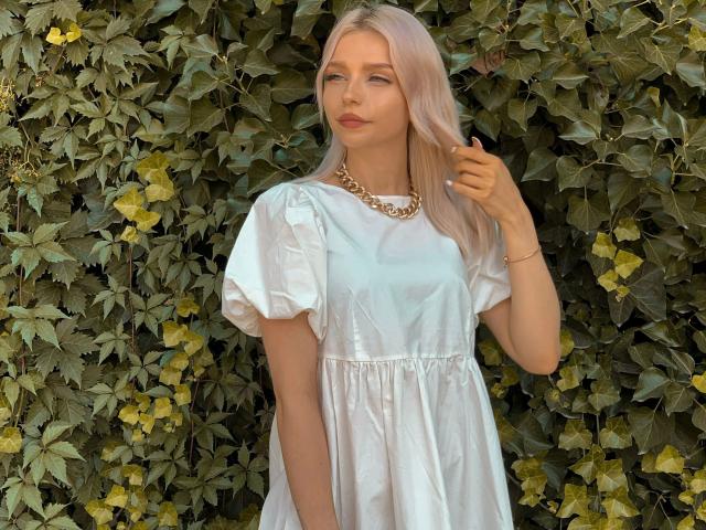 BlueCherry, mit platinblondem Haar, trägt ein weißes Kleid und eine goldene Kette und steht vor grünen Ranken, wobei sie ihre Hand an ihrem Haar berührt. Sie hat ein hübsches Gesicht und einen schönen Hintern..