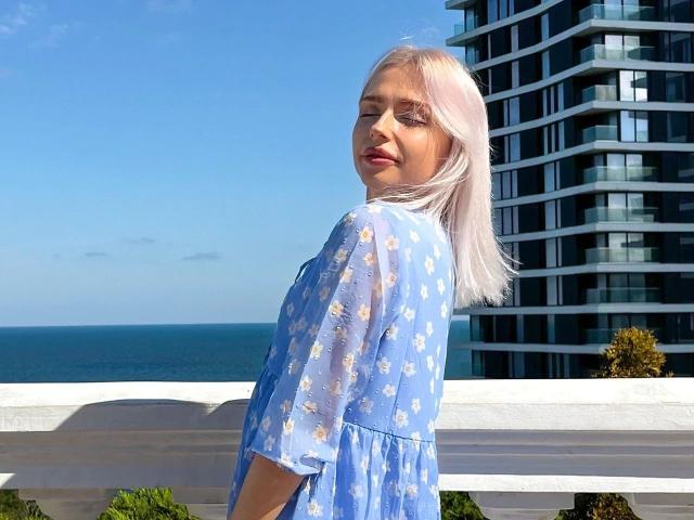 BlueCherry-hot with pale pink hair, wearing a blue floral dress, standing by a white railing, looking back with a slight smile, near a modern building and clear blue sky.