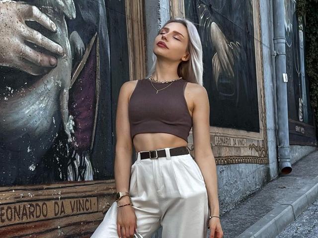 BlueCherry, attraente con pantaloni bianchi, top corto marrone e cintura, in piedi di fronte a un muro con graffiti, occhi chiusi, labbra leggermente dischiuse, indossa una collana e un braccialetto dorati, aspetto sexy..