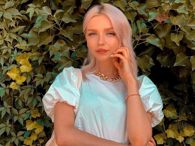 BlueCherry, com cabelo rosa, vestindo um vestido branco, colar e pulseira dourados, tocando o queixo, com um sorriso discreto, em frente a um fundo verde com folhagens..