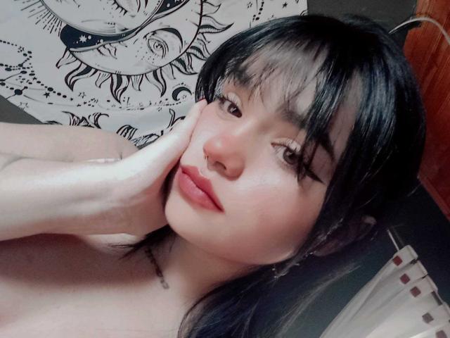 LovelyLilith with black hair, nose ring, and red lips, touching her cheek, topless, showing her smooth, pale skin and subtle makeup. Her hand rests near her face.