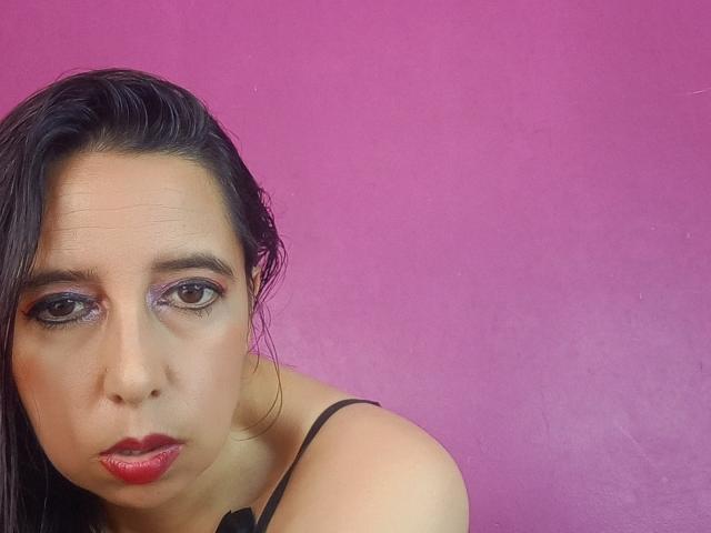 SublimeMature has dark, wet hair, purple eye makeup, red lipstick, and a serious expression, looking slightly down with black strap visible on pink background.