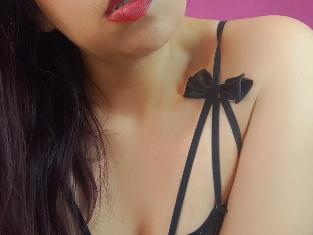 SublimeMature's upper chest with black strappy lingerie, featuring a small black bow on the right side, and her red lipstick, showing part of her black hair.