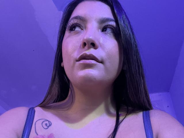 EmilyVi with long brown hair, looking up, blue-tinted lighting, visible tattoo on left shoulder, wearing a gray tank top, slight smile, bare shoulders, subtle makeup.