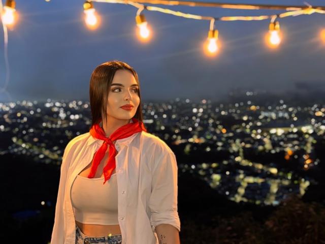 NovaDixon with medium tits, wearing a white shirt, red scarf, and white crop top, standing under string lights, looking up, city lights in the background, night time.