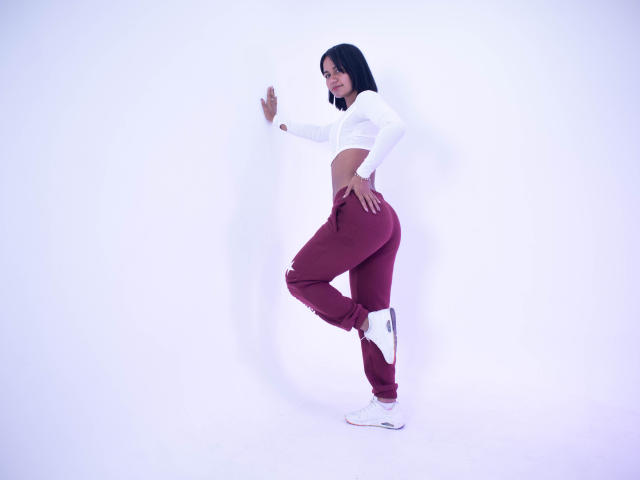 Kataleya Nava, avec des cheveux noirs, un débardeur blanc, un pantalon bordeaux, des baskets blanches, debout de côté, touchant un mur blanc, souriante, montrant ses fesses, à mi-pas, fond blanc..