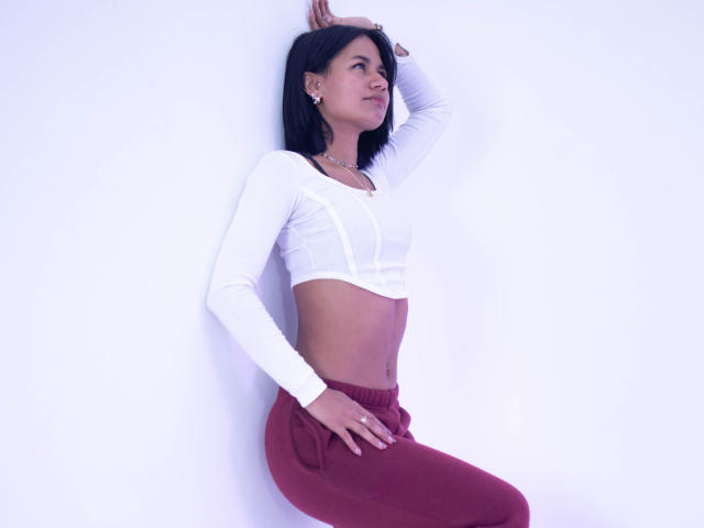 Kataleya Nava, com cabelo preto, blusa cropped branca de manga comprida, mostrando seus músculos abdominais definidos, e calças vinho, apoiada em uma parede branca, com a mão na cintura, olhando para cima..