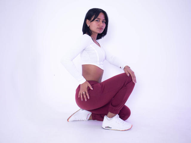 Kataleya Nava, con cabello negro, una camiseta blanca de manga larga y corte alto, pantalones rojos y zapatillas blancas, está en posición de sentadilla, con las manos en las rodillas, mostrando su trasero, sobre un fondo blanco..