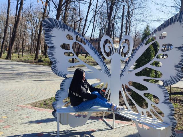 XCrazyBabeX sitting on a white butterfly-shaped bench, wearing a black jacket, blue jeans, and blue boots, with large butterfly wings behind her, in a sunny park.