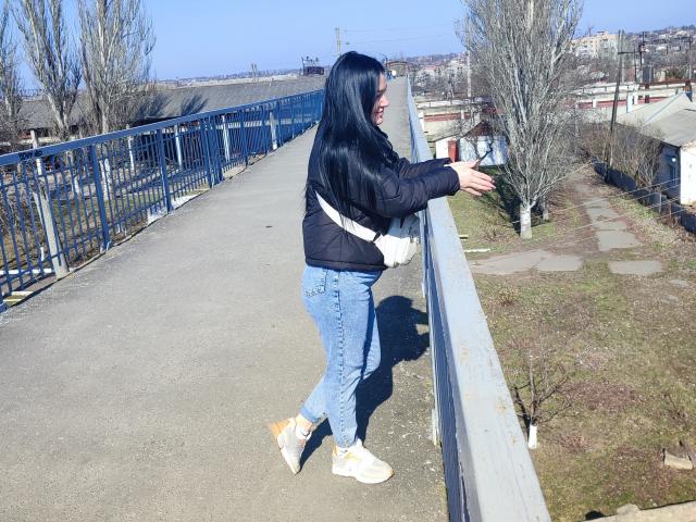 XCrazyBabeX with long black hair, black jacket, blue jeans, white shoes, and a white strap, leans on a blue railing, looking to the right.