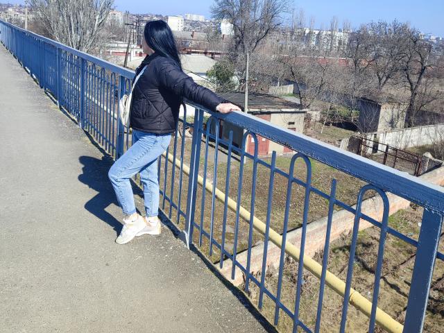 XCrazyBabeX leans against a blue metal railing, wearing a black puffer jacket, blue jeans, white sneakers, and a white crossbody bag. Bare trees and a cityscape are visible.