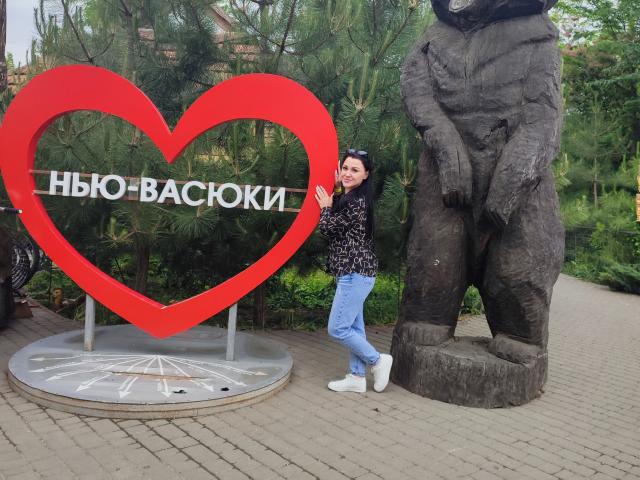 XCrazyBabeX è in piedi accanto a una statua di un orso gigante e a una scultura a forma di cuore rosso, indossa una maglietta nera con un motivo, jeans blu e scarpe da ginnastica bianche, in un parco..
