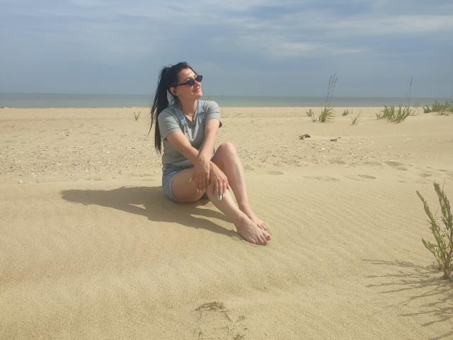 XCrazyBabeX sitting on sandy beach, wearing grey shirt and shorts, black sunglasses, smiling, long black hair, bare feet, hands on knees, small plants in background, clear blue sky.