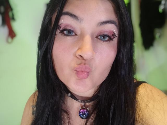 LindaSophieX with black hair, purple eyeshadow, and a nose ring, pouting her pink lips, wearing a black choker with a purple gem, blue eyes, and dark eyeliner.