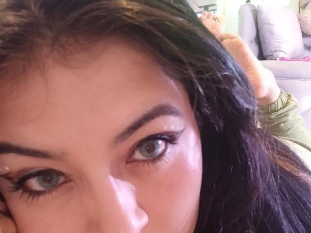 LindaSophieX's face, showing her big blue eyes with long eyelashes, dark brown eyebrows, and wet-looking skin. Her black hair is partially visible. Her green shirt is in the background.