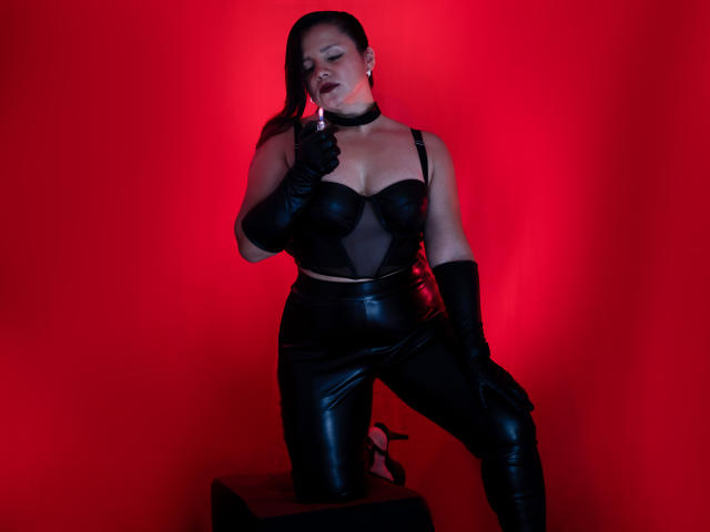 SalomeJanel with long black hair, lying on a bed, wearing black lingerie and stockings, with her tits up and ass down, looking at the camera, in a dim room.
