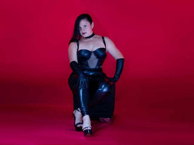 SalomeJanel with dark hair, black leather outfit, gloves, and high heels, sitting on a black cube, red background, showing her tits and ass, looking at the camera.