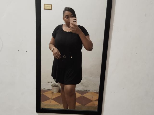 SarahSmity in a black dress, holding a pink phone, mirror selfie, showing her tits, curvy, brown skin, short black hair, wearing a watch, standing on patterned floor.