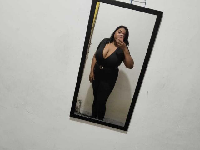 SarahSmity with large boobs, wearing a black dress with a deep cleavage, taking a mirror selfie, standing slightly bent forward, holding her phone in her right hand.