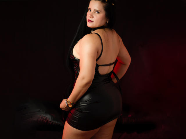 SalomeJanel with black leather dress, back to camera, showing her ass, looking over shoulder, red lipstick, black hair, bracelet, dark background, red light on right.