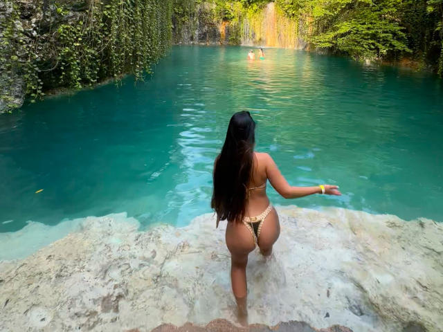 MeganDaves with long black hair, tan skin, wearing a leopard print thong, standing in clear turquoise water, facing away, showing her ass, with rocks at the bottom.
