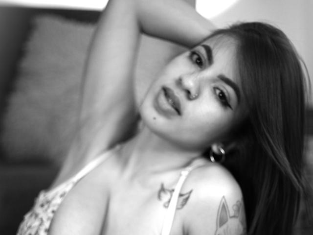 EmyBecker with her arm raised, showing tattoos on her shoulder, wearing a floral top, and looking at the camera with a slightly open mouth. Black and white photo.