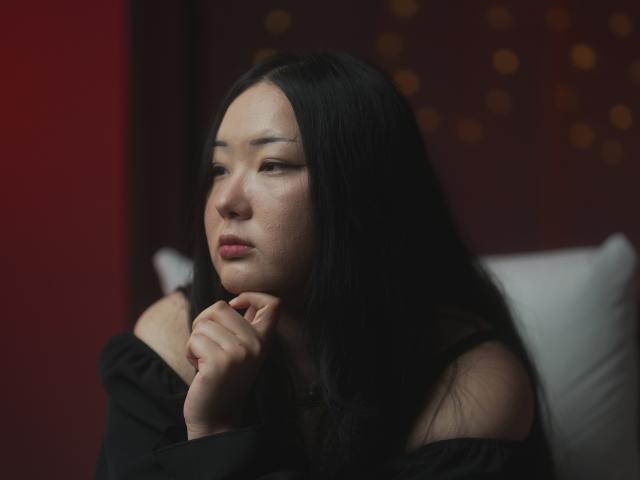 Netta, avec de longs cheveux noirs, une expression pensive, les lèvres légèrement entrouvertes, portant un haut noir à une épaule, appuyant son menton sur sa main, regardant sur le côté dans une pièce sombre..