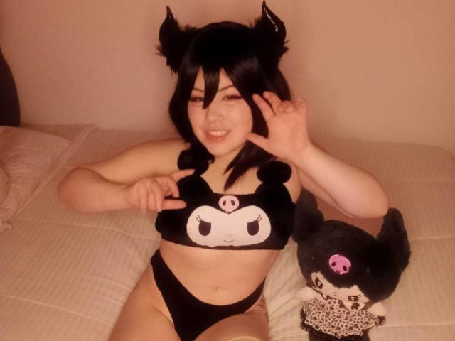 HoneeyBonny with black hair and cat ears, wearing a black bikini with a skull face, making a peace sign, and a small black stuffed cat with pink eyes.