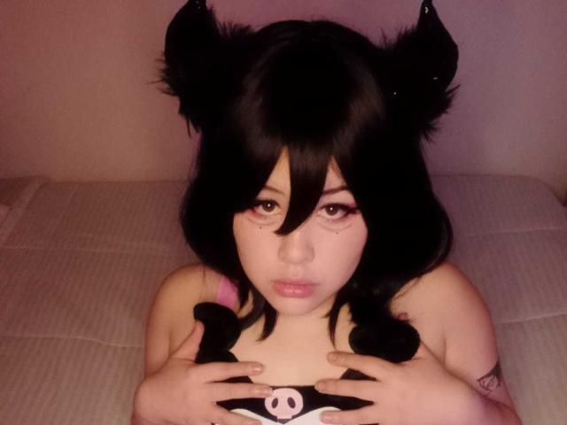 HoneeyBonny with black furry cat ears, pouty pink lips, and black hair, holding her small tits over a skull-themed top. She has a tattoo on her right arm.