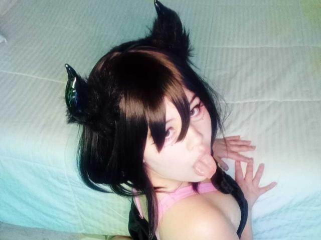 HoneeyBonny with black cat ears, sticking out her tongue, wearing a pink top, black hair with bangs, and looking up at the camera. Pale skin, white bed in background.