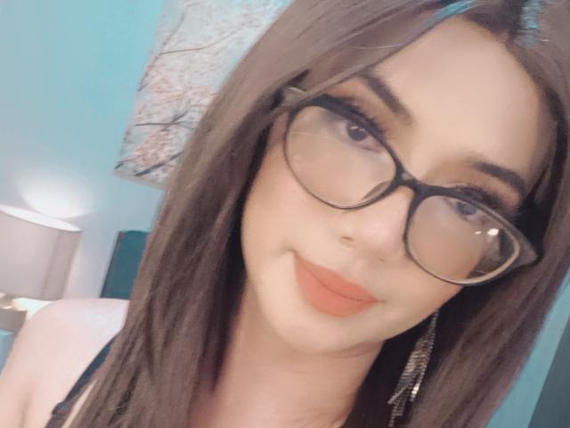 TransKathie with glasses, brown hair, and orange lipstick, showing a slight smile, with visible tits and a cute face. She has a light skin tone and looks attractive.