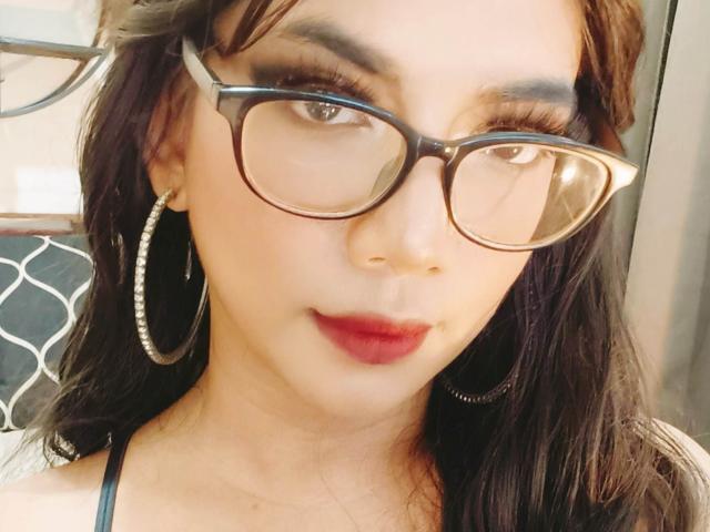 TransKathie has dark brown hair, glasses, red lipstick, hoop earrings, and a slightly smiling face with light skin and brown eyes. Boobs not visible. Soft, warm lighting.