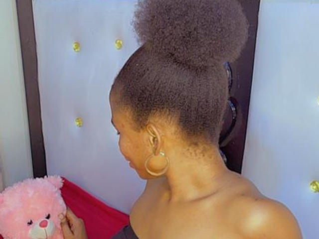 SammyBea with a high, curly afro, holding a pink fluffy toy, wearing gold hoop earrings, and a gray off-shoulder top, showing her bare shoulder and neck.