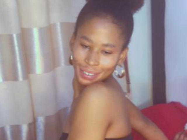 SammyBea with dark skin, brown hair in a bun, wearing earrings, smiling, showing bare shoulders, and red fabric in the background. Boobs partially visible. Soft, warm lighting.