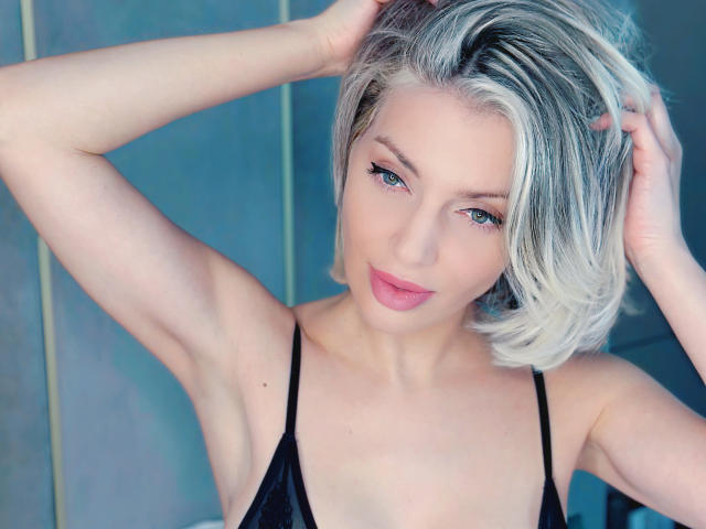 AmmyMadison con capelli biondi, occhi azzurri, labbra rosa, top nero a spalline, braccia alzate, mani tra i capelli, sorridente, luce soffusa, pelle chiara, seno di medie dimensioni, trucco leggero, posa informale..