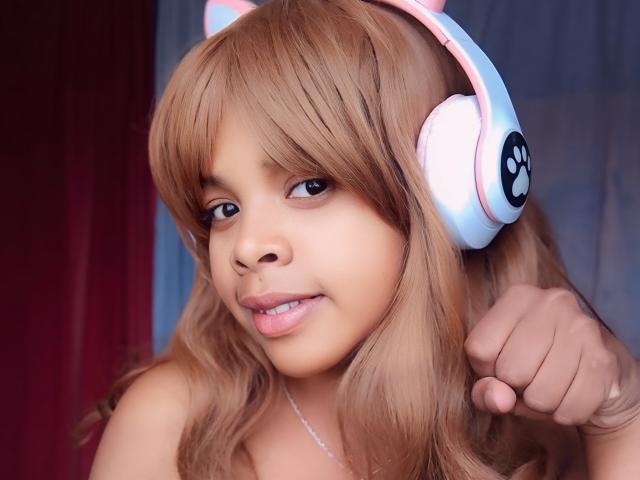 FireGirlGorgeux with light brown hair, wearing pink and white cat-ear headphones, has cute pink lips and a small nose, her hand near her face, looking slightly to the side.