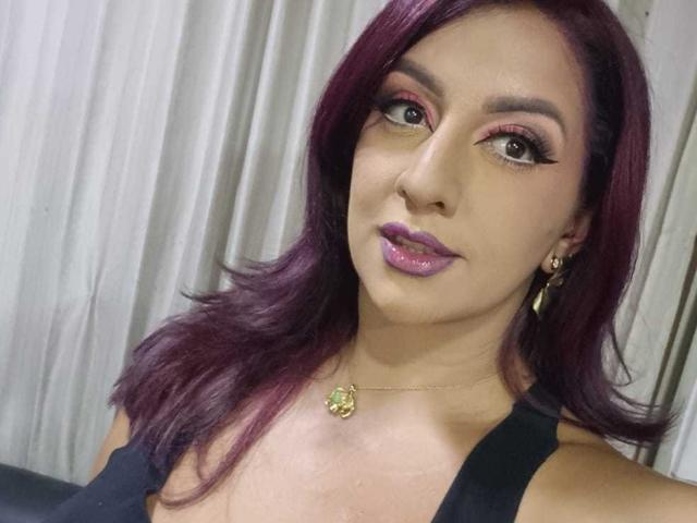 AndreaSharick has purple lipstick, dark eye makeup, and long purple hair. She's wearing a black top, gold necklace with a green flower, and gold earrings.