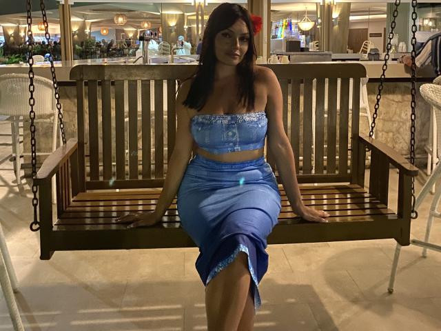 GalaMalkova sits on a wooden swing, wearing a blue strapless top and matching skirt, showing her tits and ass. She has dark hair and a red flower in her hair.